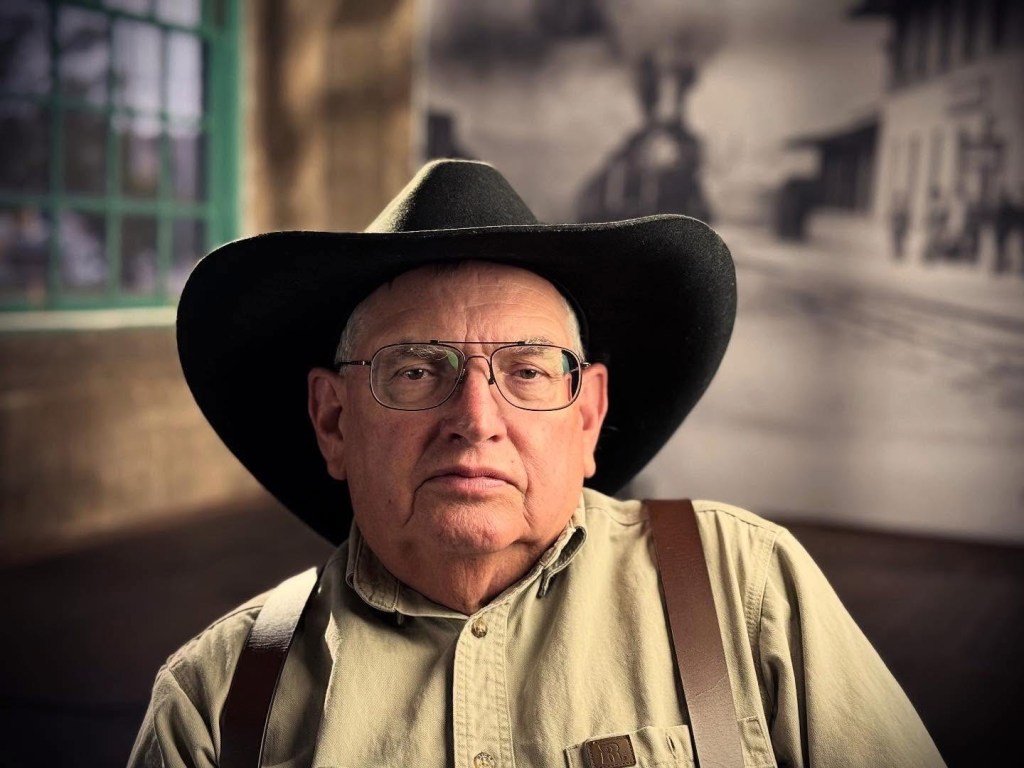 A senior man wearing a large black hat and glasses, dressed in a button-up shirt with suspenders, sitting indoors with a vintage train station backdrop.
