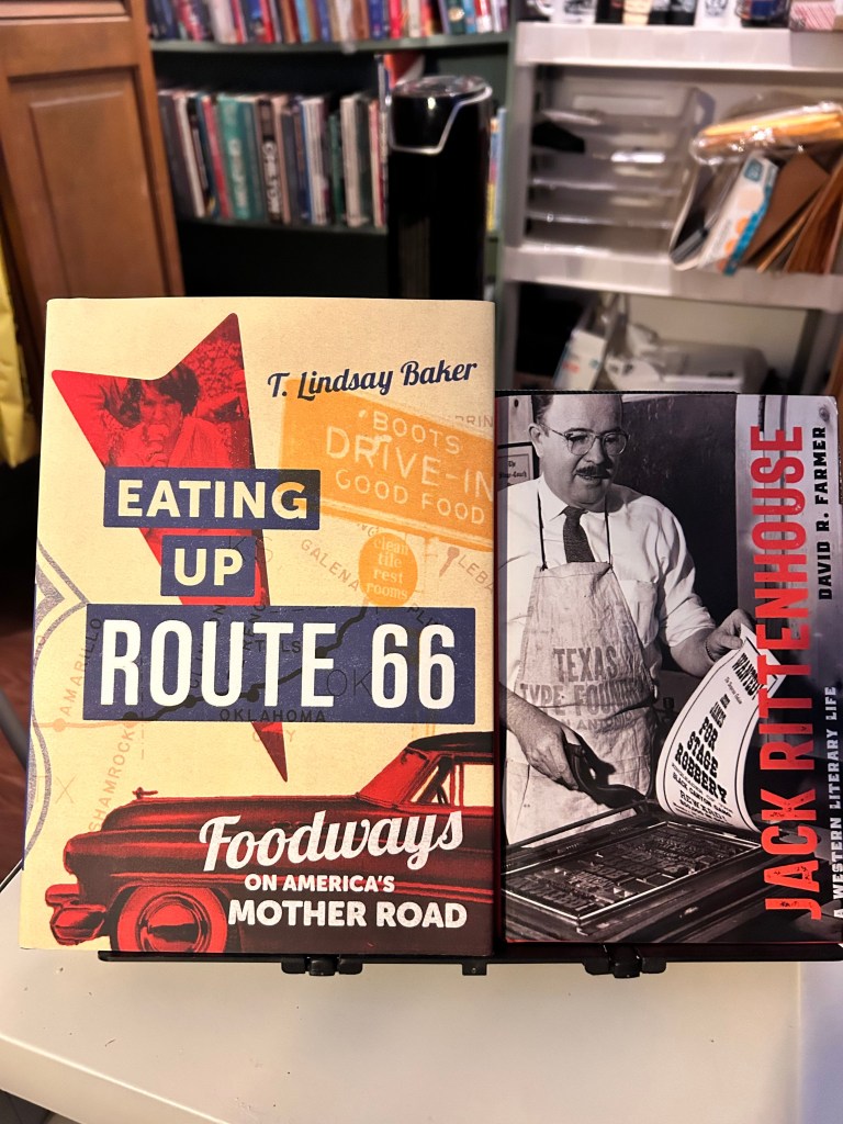 Two books displayed side by side on a table. The left book titled 'Eating Up Route 66' by T. Lindsay Baker, featuring a vintage design with red accents and images related to Route 66. The right book titled 'Jack Rittenhouse' by David R. Farmer, showcasing a black and white photo of a man in an apron, surrounded by typewriter elements. Background includes shelves of books.